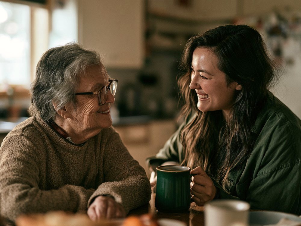 A senior and her companion talking over mugs of tea at a kitchen table